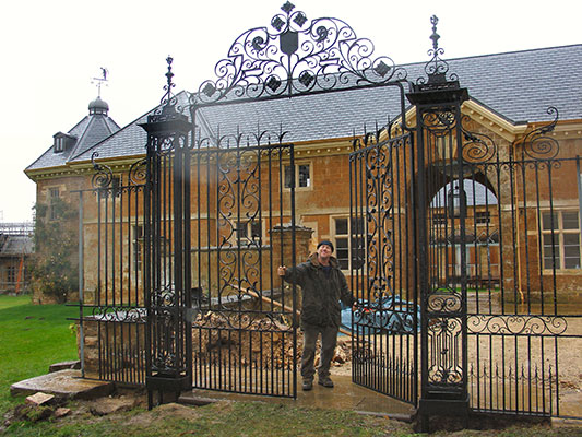 The almost completion of a particularly large and ornate pair of gates with iron supporting columns and a large ornate arch containing the family crest spanning over these.